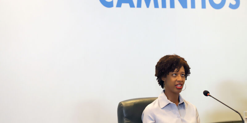 Uma mulher negra está sentada atrás de uma mesa de madeira em um ambiente que parece um auditório ou sala de palestras. Ela tem cabelos curtos e cacheados, usa camisa social clara e brincos grandes de argola. À sua frente há um microfone ligado, indicando que ela está falando para um público. Ela está sentada em uma cadeira preta e olha levemente para cima enquanto fala, com expressão séria e atenta. Sobre a mesa há alguns objetos: um celular, um teclado e uma pequena tigela preta. Ao fundo existe uma grande tela branca de projeção. Na parte superior da tela aparece a palavra “CAMINHOS” escrita em letras grandes azuis.