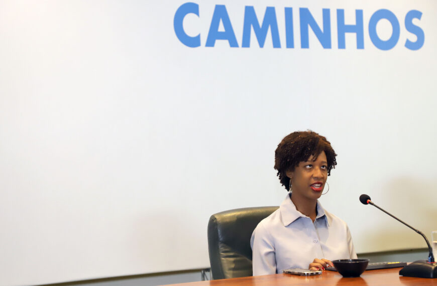 Uma mulher negra está sentada atrás de uma mesa de madeira em um ambiente que parece um auditório ou sala de palestras. Ela tem cabelos curtos e cacheados, usa camisa social clara e brincos grandes de argola. À sua frente há um microfone ligado, indicando que ela está falando para um público. Ela está sentada em uma cadeira preta e olha levemente para cima enquanto fala, com expressão séria e atenta. Sobre a mesa há alguns objetos: um celular, um teclado e uma pequena tigela preta. Ao fundo existe uma grande tela branca de projeção. Na parte superior da tela aparece a palavra “CAMINHOS” escrita em letras grandes azuis.