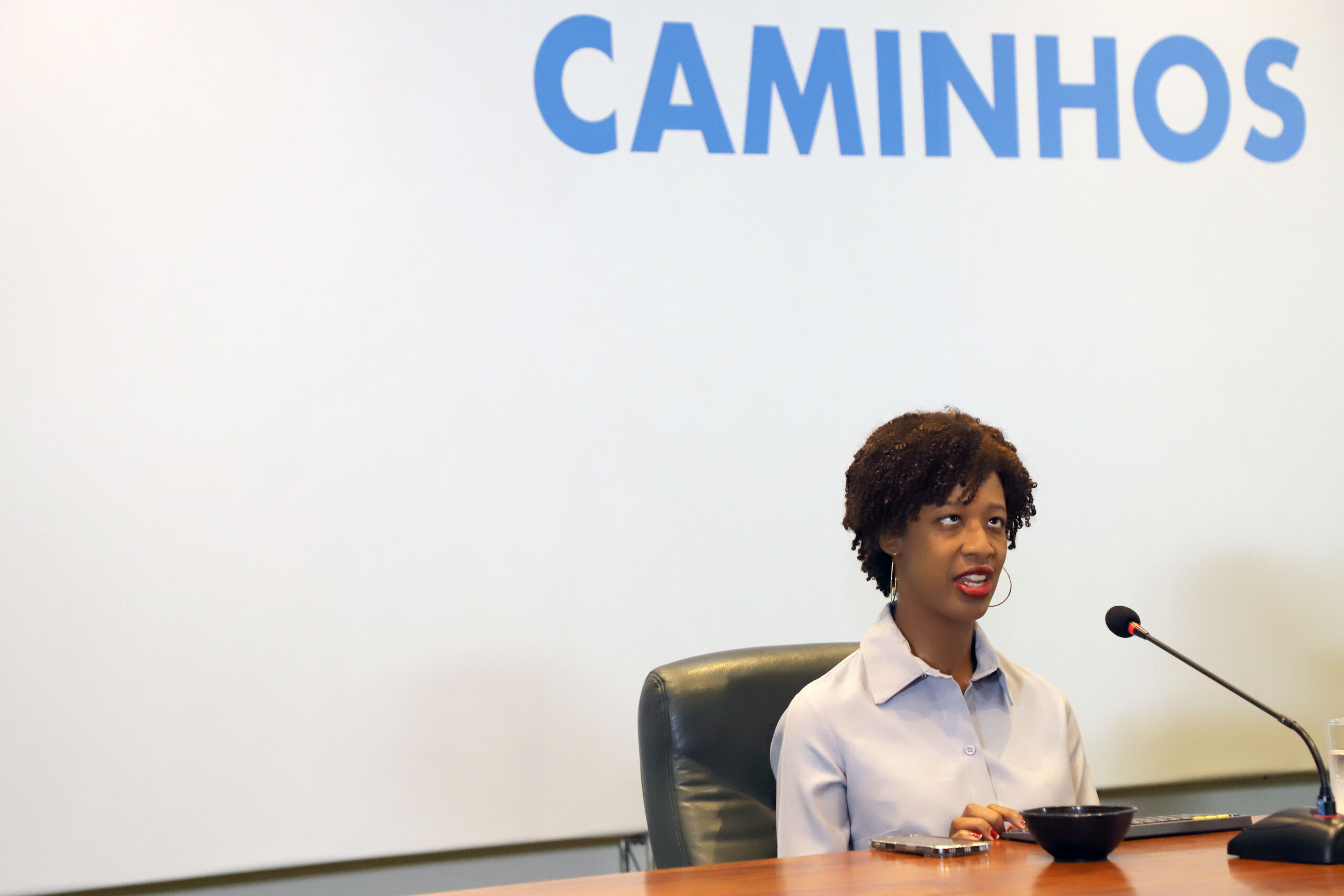 Uma mulher negra está sentada atrás de uma mesa de madeira em um ambiente que parece um auditório ou sala de palestras. Ela tem cabelos curtos e cacheados, usa camisa social clara e brincos grandes de argola. À sua frente há um microfone ligado, indicando que ela está falando para um público. Ela está sentada em uma cadeira preta e olha levemente para cima enquanto fala, com expressão séria e atenta. Sobre a mesa há alguns objetos: um celular, um teclado e uma pequena tigela preta. Ao fundo existe uma grande tela branca de projeção. Na parte superior da tela aparece a palavra “CAMINHOS” escrita em letras grandes azuis.
