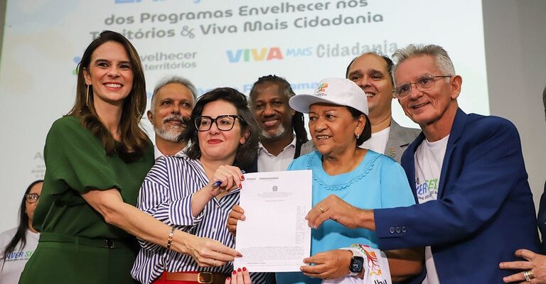 Em um palco de evento institucional, sete pessoas posam juntas sorrindo para uma foto durante a apresentação de um documento oficial. Ao centro da imagem, duas mulheres seguram um papel com aparência de ofício ou termo institucional, enquanto as demais pessoas ao redor participam do momento de entrega simbólica. À esquerda, uma mulher de vestido verde escuro sorri para a câmera. Ao lado dela, uma mulher de óculos, usando camisa listrada em branco e azul e calça vermelha, segura o documento. No centro, uma mulher de blusa azul-clara e boné branco também segura o papel e sorri. Ao redor delas, há homens e mulheres vestidos de maneira informal e institucional, alguns usando camisetas relacionadas ao evento. À direita, um homem de blazer azul e camiseta branca posa sorrindo. No fundo do palco, um telão exibe o texto “Lançamento dos Programas Envelhecer nos Territórios & Viva Mais Cidadania”, além das logomarcas dos programas e do governo.