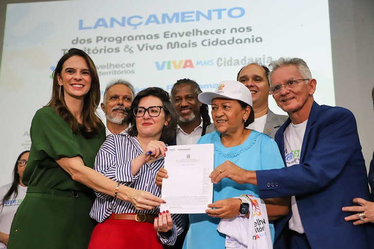 Em um palco de evento institucional, sete pessoas posam juntas sorrindo para uma foto durante a apresentação de um documento oficial. Ao centro da imagem, duas mulheres seguram um papel com aparência de ofício ou termo institucional, enquanto as demais pessoas ao redor participam do momento de entrega simbólica. À esquerda, uma mulher de vestido verde escuro sorri para a câmera. Ao lado dela, uma mulher de óculos, usando camisa listrada em branco e azul e calça vermelha, segura o documento. No centro, uma mulher de blusa azul-clara e boné branco também segura o papel e sorri. Ao redor delas, há homens e mulheres vestidos de maneira informal e institucional, alguns usando camisetas relacionadas ao evento. À direita, um homem de blazer azul e camiseta branca posa sorrindo. No fundo do palco, um telão exibe o texto “Lançamento dos Programas Envelhecer nos Territórios & Viva Mais Cidadania”, além das logomarcas dos programas e do governo.