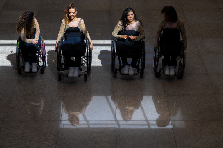 Quatro mulheres usuárias de cadeira de rodas estão alinhadas lado a lado em um ambiente interno amplo, com iluminação dramática e reflexos no piso brilhante. Todas usam roupas semelhantes: blusa preta, tecido transparente com brilho dourado nos braços e tênis brancos. As cadeiras de rodas são diferentes entre si, algumas com detalhes coloridos nas rodas. A iluminação cria um forte contraste entre luz e sombra. As duas mulheres ao centro estão mais iluminadas, enquanto as das extremidades aparecem parcialmente na sombra. Cada uma demonstra uma expressão e postura diferente: uma olha para baixo, outra sorri suavemente, uma terceira mantém expressão séria e concentrada, e a quarta aparece mais escurecida pela sombra. O piso polido reflete as mulheres e as cadeiras de rodas, criando um efeito visual elegante e artístico.