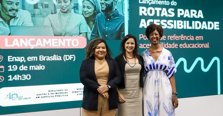 Três mulheres posam em frente a um grande painel digital de divulgação do evento “Lançamento do Rotas para Acessibilidade”. O painel, em tons de verde e azul, informa que o evento ocorreu na Enap, em Brasília (DF), no dia 19 de maio, às 14h30, e destaca a frase “Portal de referência em acessibilidade educacional”. No centro da imagem, as três mulheres estão em pé, lado a lado, sorrindo para a câmera. À esquerda, uma mulher de cabelos cacheados usa conjunto bege e blazer azul-escuro. No meio, uma mulher de cabelos escuros e lisos veste um vestido bege claro. À direita, uma mulher negra, de cabelos curtos e volumosos, usa um vestido branco com estampas azuis.
