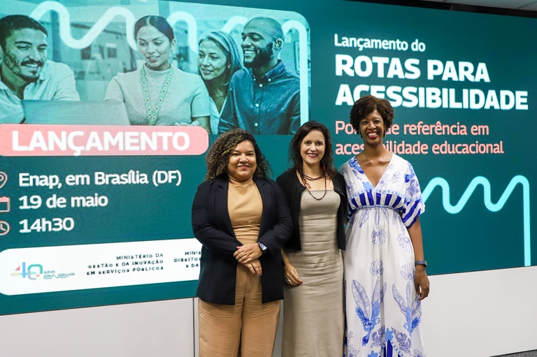 Três mulheres posam em frente a um grande painel digital de divulgação do evento “Lançamento do Rotas para Acessibilidade”. O painel, em tons de verde e azul, informa que o evento ocorreu na Enap, em Brasília (DF), no dia 19 de maio, às 14h30, e destaca a frase “Portal de referência em acessibilidade educacional”. No centro da imagem, as três mulheres estão em pé, lado a lado, sorrindo para a câmera. À esquerda, uma mulher de cabelos cacheados usa conjunto bege e blazer azul-escuro. No meio, uma mulher de cabelos escuros e lisos veste um vestido bege claro. À direita, uma mulher negra, de cabelos curtos e volumosos, usa um vestido branco com estampas azuis.