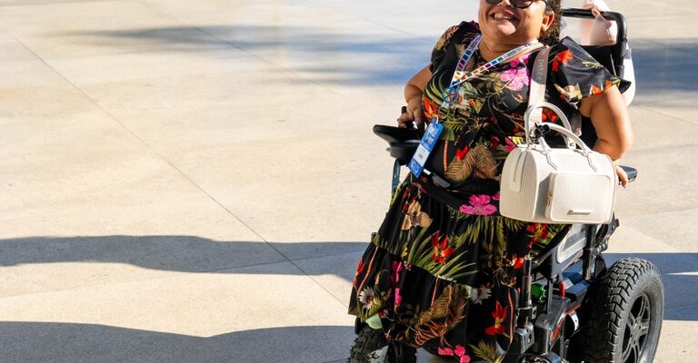 Uma mulher sorridente está em uma cadeira de rodas motorizada sobre um piso amplo e claro, em uma área externa iluminada pelo sol. Ela usa óculos escuros e um vestido preto estampado com flores coloridas e folhagens tropicais em tons vibrantes, como rosa, vermelho, verde e amarelo. A mulher está posicionada no lado direito da imagem e aparenta estar feliz e confiante. Em seu colo há uma bolsa branca estruturada. No pescoço, ela usa um crachá com fitas coloridas, sugerindo participação em um evento ou atividade institucional. A cadeira de rodas possui rodas grandes e robustas, adequadas para diferentes tipos de terreno. A luz do sol cria sombras longas no chão, trazendo sensação de fim de tarde ou início da manhã.