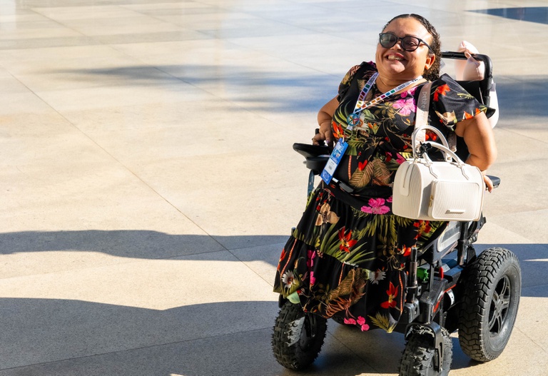 Uma mulher sorridente está em uma cadeira de rodas motorizada sobre um piso amplo e claro, em uma área externa iluminada pelo sol. Ela usa óculos escuros e um vestido preto estampado com flores coloridas e folhagens tropicais em tons vibrantes, como rosa, vermelho, verde e amarelo. A mulher está posicionada no lado direito da imagem e aparenta estar feliz e confiante. Em seu colo há uma bolsa branca estruturada. No pescoço, ela usa um crachá com fitas coloridas, sugerindo participação em um evento ou atividade institucional. A cadeira de rodas possui rodas grandes e robustas, adequadas para diferentes tipos de terreno. A luz do sol cria sombras longas no chão, trazendo sensação de fim de tarde ou início da manhã.