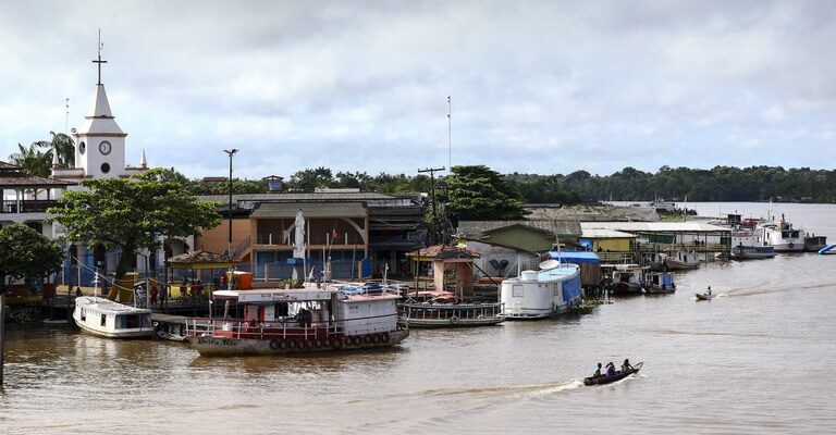 Comunidade ribeirinha com barcos atracados às margens de um rio largo, igreja com torre branca e céu encoberto por nuvens.