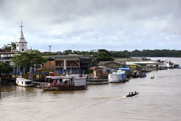 Comunidade ribeirinha com barcos atracados às margens de um rio largo, igreja com torre branca e céu encoberto por nuvens.