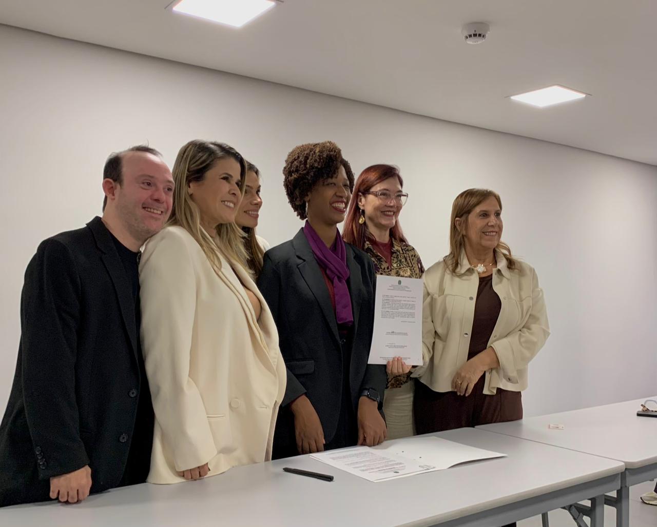 NVSL Seis pessoas estão em pé, lado a lado, voltadas para a câmera. Entre elas, há um homem e cinco mulheres. O grupo está posicionado atrás de uma mesa clara. Da esquerda para a direita, aparece um homem com cabelo curto usando roupa escura. Ao lado dele, uma mulher veste blazer claro. Em seguida, outra mulher aparece parcialmente visível atrás das demais. No centro, uma mulher com cabelo curto e cacheado usa blazer escuro e lenço roxo. Ao seu lado, uma mulher de cabelos avermelhados, usando óculos e roupa estampada, segura uma folha de papel impressa. À direita, uma mulher veste casaco claro sobre roupa escura. No teto, são visíveis luminárias retangulares e um detector de fumaça. O ambiente tem aparência de sala de reunião ou espaço institucional.