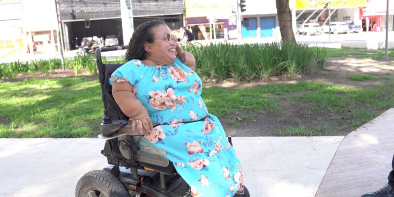 Mulher usando cadeira de rodas motorizada conversa ao ar livre em área com gramado e árvores.