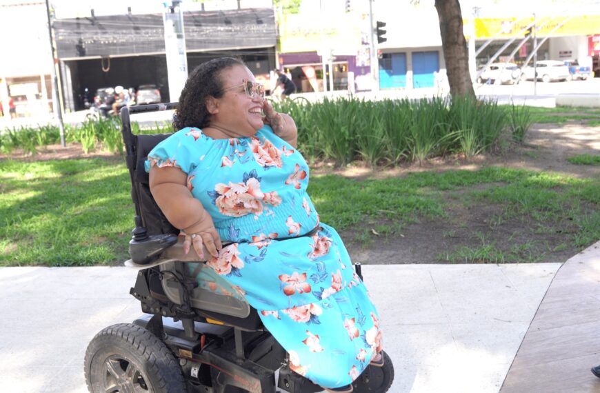 Mulher usando cadeira de rodas motorizada conversa ao ar livre em área com gramado e árvores.