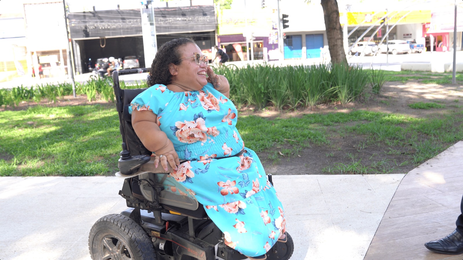 Mulher usando cadeira de rodas motorizada conversa ao ar livre em área com gramado e árvores.