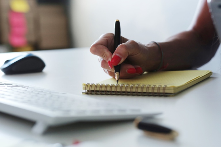 Mão com unhas pintadas de vermelho escrevendo com uma caneta preta em um bloco de notas amarelo sobre uma mesa de escritório.