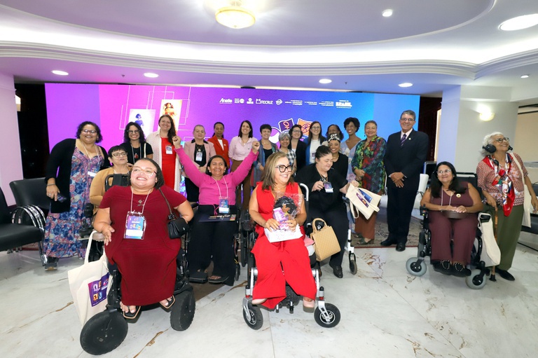 Grupo diverso de pessoas, incluindo várias mulheres usuárias de cadeira de rodas, sorriem e comemoram juntas durante evento oficial em palco com painel institucional ao fundo.