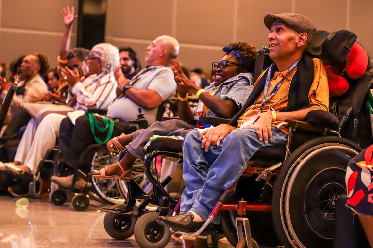 Pessoas em cadeiras de rodas e demais participantes da plateia sorriem e aplaudem durante evento em auditório.